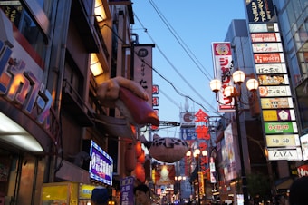 A bustling urban street filled with bright, colorful neon signs and advertisements in Japanese characters. Large, visually striking models such as a giant sushi and pufferfish hang outside storefronts. The street is crowded, with many people visible in the foreground. Electric wires crisscross overhead, and the buildings are closely packed together, contributing to a vibrant, energetic atmosphere.
