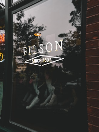 A storefront window with the word 'Filson' and 'Since 1897' in bold white letters. The window reflects trees, a building, and a street scene outside. Inside the store, several plush stuffed animals resembling large dogs are visible.