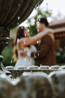 A romantic scene focusing on a stone fountain with water droplets falling from it. In the blurred background, a couple can be seen embracing, dressed in formal attire.