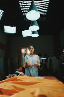 A person stands in a dimly lit room, which appears to be a creative workspace or studio. Bright overhead lights illuminate a table covered with yellow fabric. The individual holds a pencil, wearing a casual, colorful shirt. In the background, several people are engaged in activities related to textile or garment making.