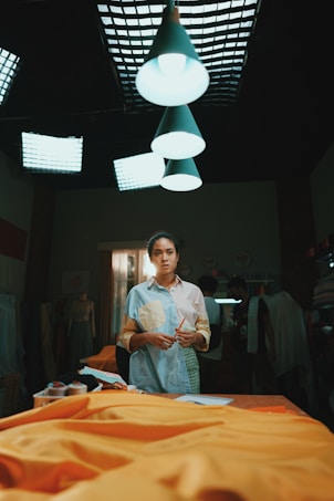 A person stands in a dimly lit room, which appears to be a creative workspace or studio. Bright overhead lights illuminate a table covered with yellow fabric. The individual holds a pencil, wearing a casual, colorful shirt. In the background, several people are engaged in activities related to textile or garment making.