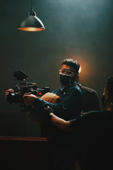 A person wearing a mask is operating a professional video camera under dim lighting. The scene is lit by a hanging industrial-style lamp, creating a focused and moody atmosphere. Another person is partially visible to the side, assisting or observing.