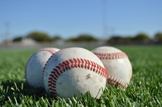 Three baseballs are positioned closely together on a lush green grass field, suggesting a sports setting. The focus is on the baseballs, which have visible stitching and slight wear, indicating use. The background is blurred, with hints of a clear blue sky and a distant landscape.
