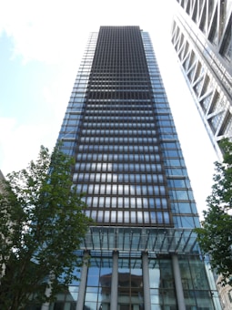 A tall, modern skyscraper with reflective glass panels stretches into the sky. The architecture appears sleek and contemporary, with a grid-like pattern on its facade. Trees with green leaves are visible in the foreground, framing the building. Adjacent structures are visible with a similar modern design.