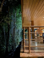 A cozy wooden deck is adjacent to a dense bamboo forest. The scene is warmly lit, with bamboo chairs arranged on the tiled floor and wooden railings lining the edge.