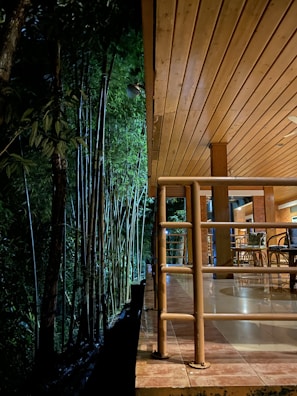 A cozy wooden deck is adjacent to a dense bamboo forest. The scene is warmly lit, with bamboo chairs arranged on the tiled floor and wooden railings lining the edge.
