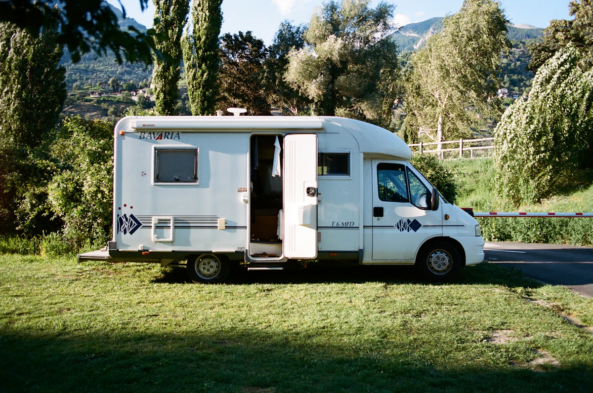 Un camping-car blanc à la porte ouverte, stationné sur un emplacement herbeux bordé d'arbres, avec des montagnes en arrière-plan