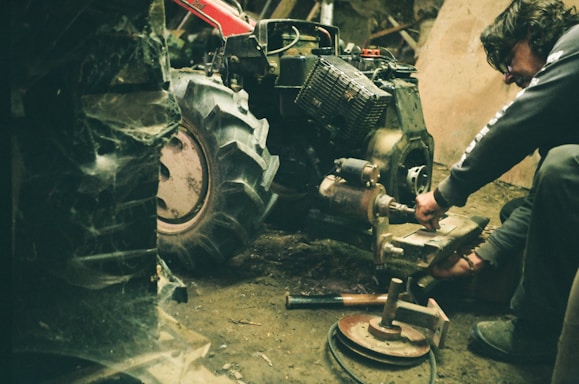 A person working on a large piece of machinery in a dimly lit, cluttered space. The machinery appears to be a tractor or another type of heavy equipment. The environment looks industrial and has a rustic, gritty feel with visible dirt and spider webs.