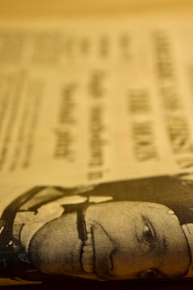 A close-up of a newspaper page with printed text and a low-resolution image of a man's face. The image is in a sepia tone, giving it a vintage look.