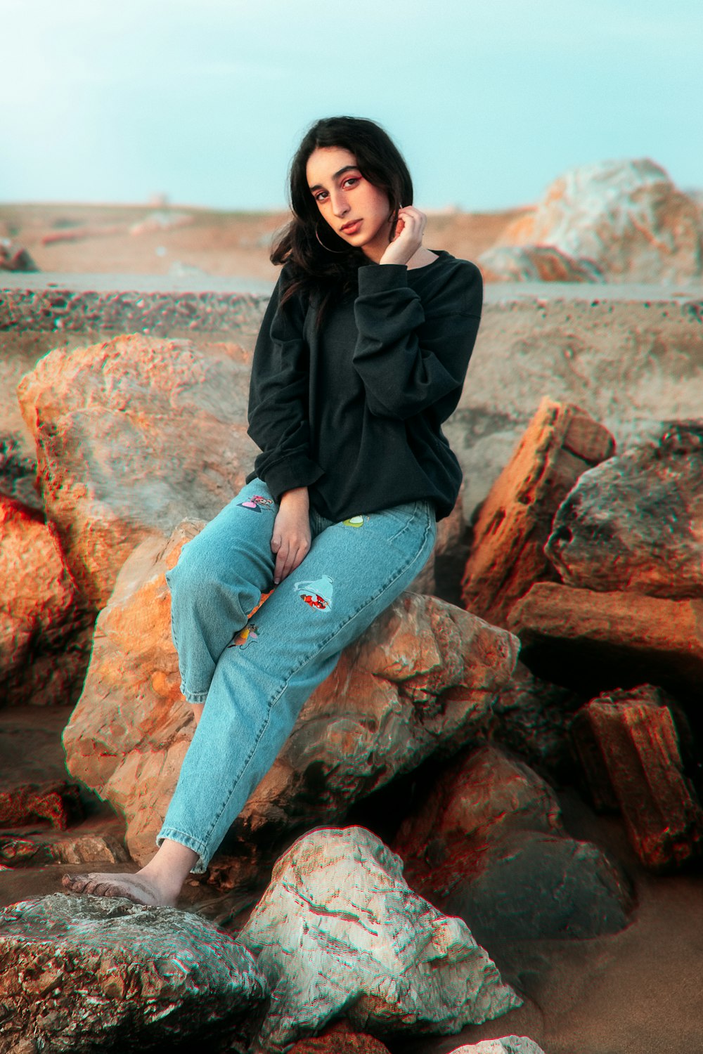woman in black jacket and blue denim jeans sitting on brown rock during daytime