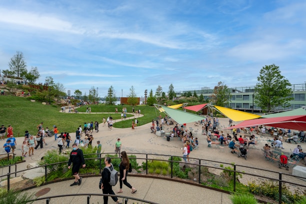 A lively outdoor public space with people walking, sitting, and socializing. There are colorful triangular shade structures in red, yellow, and green. The area is surrounded by landscaped grass and trees, and people are enjoying a pleasant day.