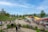 A lively outdoor public space with people walking, sitting, and socializing. There are colorful triangular shade structures in red, yellow, and green. The area is surrounded by landscaped grass and trees, and people are enjoying a pleasant day.