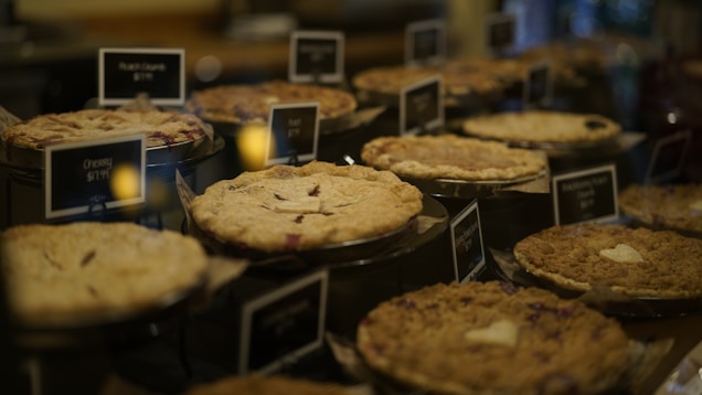 A variety of pies are arranged in a display case, each with a small label indicating the type and price. The pies have different crust styles and fillings, creating a diverse and appetizing arrangement.