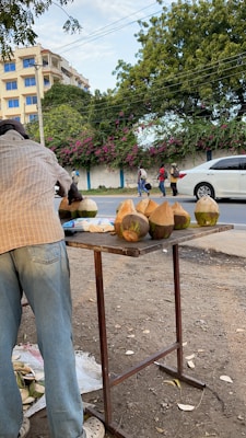 A street vendor is preparing fresh coconuts on a wooden table by the roadside. There are several coconuts with their tops cut off, ready to be served. In the background, a few people are walking along the sidewalk, and a white car is passing by. The street is lined with a wall covered in flowering plants, and there's a large tree overhead.