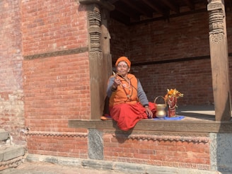 A person dressed in traditional attire is sitting on a platform in front of an intricately carved wooden structure. They hold a gesture with one hand and are surrounded by a brass pot, flowers, and a cloth. The background is a wall made of reddish bricks.