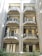 A multi-story residential building with six balconies featuring geometric metal railings and pale-colored walls. The balconies display various items including potted plants, furniture, and air conditioning units. Windows with shutters are open, allowing a glimpse into the interiors. Vegetation can be seen at the base of the building.