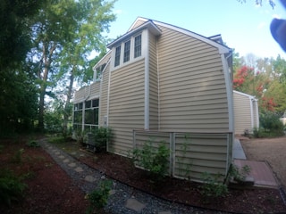 The image depicts a side view of a house with light brown siding, surrounded by trees and greenery. A stone pathway leads through a garden area, with shrubs and plants lining the path. The house features multiple windows and a screened porch area.
