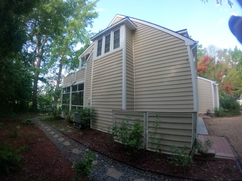 The image depicts a side view of a house with light brown siding, surrounded by trees and greenery. A stone pathway leads through a garden area, with shrubs and plants lining the path. The house features multiple windows and a screened porch area.