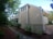 The image depicts a side view of a house with light brown siding, surrounded by trees and greenery. A stone pathway leads through a garden area, with shrubs and plants lining the path. The house features multiple windows and a screened porch area.