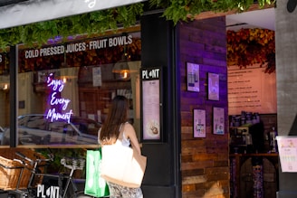 A person stands in front of a juice and smoothie shop, looking at the menu displayed on the wall. The shop's window features a neon sign with the words 'Enjoy Every Moment,' and there are decorative green plants above. A bicycle with a wicker basket is parked outside the shop.