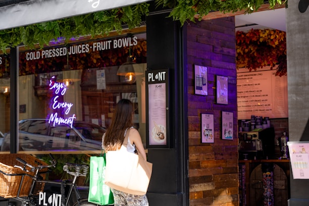 A person stands in front of a juice and smoothie shop, looking at the menu displayed on the wall. The shop's window features a neon sign with the words 'Enjoy Every Moment,' and there are decorative green plants above. A bicycle with a wicker basket is parked outside the shop.
