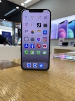 A smartphone with a black frame is displayed on a wooden surface, showcasing its home screen filled with various app icons. In the background, a retail store environment is visible with bright lighting and blurred promotional posters along the walls.