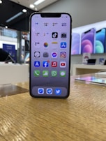 A smartphone with a black frame is displayed on a wooden surface, showcasing its home screen filled with various app icons. In the background, a retail store environment is visible with bright lighting and blurred promotional posters along the walls.