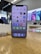 A smartphone with a black frame is displayed on a wooden surface, showcasing its home screen filled with various app icons. In the background, a retail store environment is visible with bright lighting and blurred promotional posters along the walls.