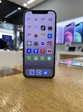 A smartphone with a black frame is displayed on a wooden surface, showcasing its home screen filled with various app icons. In the background, a retail store environment is visible with bright lighting and blurred promotional posters along the walls.