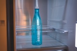 A blue bottle with the brand name Samsung written on it is placed inside a refrigerator. The interior of the fridge is mostly empty, with metal shelves visible in the background.