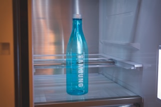 A blue bottle with the brand name Samsung written on it is placed inside a refrigerator. The interior of the fridge is mostly empty, with metal shelves visible in the background.