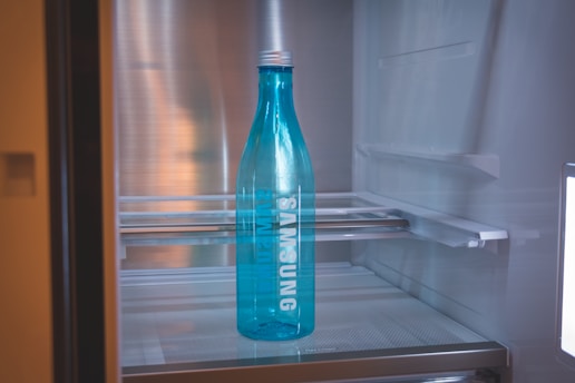 A blue bottle with the brand name Samsung written on it is placed inside a refrigerator. The interior of the fridge is mostly empty, with metal shelves visible in the background.