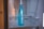 A blue bottle with the brand name Samsung written on it is placed inside a refrigerator. The interior of the fridge is mostly empty, with metal shelves visible in the background.