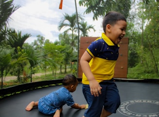 Two young children play joyfully on a trampoline surrounded by lush greenery. One child stands and laughs while the other, dressed in blue, crawls on the trampoline surface. Tall trees and a cloudy sky can be seen in the background, adding a natural, outdoor setting.