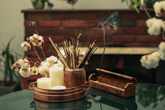 A cozy arrangement features a copper tray holding several white candles, dried cotton stems, and a decorative container filled with incense sticks. Smoke gently rises from a lit incense stick placed in a wooden holder, and the scene is set against a backdrop of a brick wall. The ambiance suggests a calm, meditative environment.