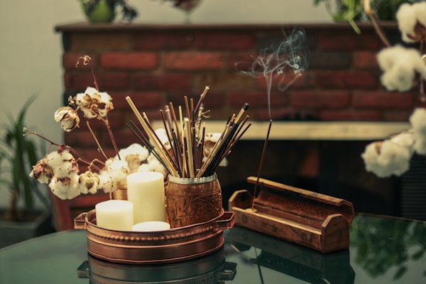 A cozy arrangement features a copper tray holding several white candles, dried cotton stems, and a decorative container filled with incense sticks. Smoke gently rises from a lit incense stick placed in a wooden holder, and the scene is set against a backdrop of a brick wall. The ambiance suggests a calm, meditative environment.