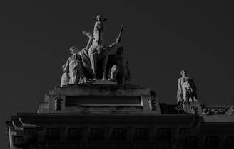 Stone sculptures adorn the top of a grand architectural structure. The central figure is a seated person with a headdress, flanked by other figures standing in classical poses. The image is in black and white, highlighting the shadows and details in the stonework.
