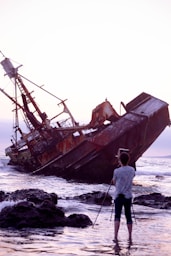 man in white shirt and gray pants standing on rocky shore near brown ship during daytime