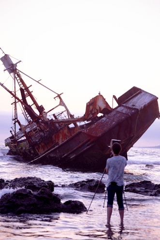 man in white shirt and gray pants standing on rocky shore near brown ship during daytime