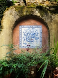 A decorative ceramic tile featuring a detailed blue floral design is mounted on an old, weathered stone wall. The tile is framed with an ornate blue border, contrasting against the earthy, moss-covered structure. Below, lush green foliage and plants in terracotta pots add a touch of nature to the rustic setting.