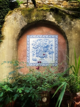 A decorative ceramic tile featuring a detailed blue floral design is mounted on an old, weathered stone wall. The tile is framed with an ornate blue border, contrasting against the earthy, moss-covered structure. Below, lush green foliage and plants in terracotta pots add a touch of nature to the rustic setting.