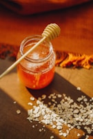 A glass jar filled with honey is placed on a wooden surface, accompanied by a honey dipper resting on top. Scattered around the jar are small piles of oats. The scene is warmly lit, casting soft shadows and highlighting the rich amber color of the honey.