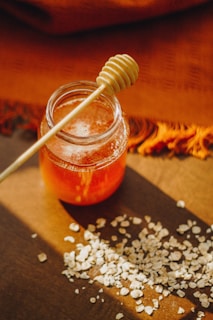 A glass jar filled with honey is placed on a wooden surface, accompanied by a honey dipper resting on top. Scattered around the jar are small piles of oats. The scene is warmly lit, casting soft shadows and highlighting the rich amber color of the honey.