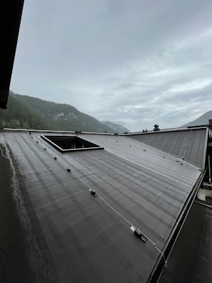 A sloped roof with a smooth, dark surface is seen with metal fixtures evenly distributed across it. In the background, mist-covered mountains and a cloudy sky create a moody atmosphere.