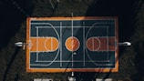 An aerial view of an outdoor basketball court with a striking rectangular layout. The court's surface features dark green and orange colors with white lines marking the playing areas. Four basketball hoops are positioned at both ends. The court is surrounded by grassy areas, and shadows cast by trees or poles intersect the court, adding dramatic contrast.