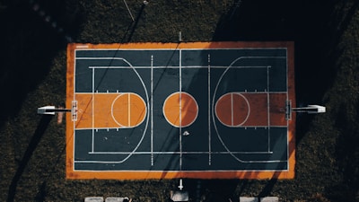An aerial view of an outdoor basketball court with a striking rectangular layout. The court's surface features dark green and orange colors with white lines marking the playing areas. Four basketball hoops are positioned at both ends. The court is surrounded by grassy areas, and shadows cast by trees or poles intersect the court, adding dramatic contrast.