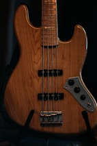 A close-up view of a wooden electric bass guitar featuring four strings and a classic pickup design. The body of the guitar displays a natural wood grain finish with a polished metallic control plate and knobs.