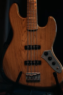 A close-up view of a wooden electric bass guitar featuring four strings and a classic pickup design. The body of the guitar displays a natural wood grain finish with a polished metallic control plate and knobs.
