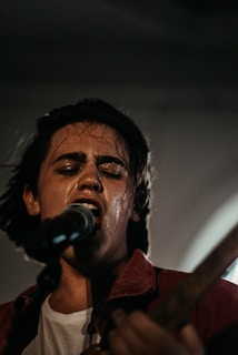 A person passionately singing into a microphone with closed eyes, implying deep emotional engagement. The setting appears dimly lit, creating a moody atmosphere. The individual is wearing a dark shirt and is slightly sweaty, suggesting a dynamic performance. The focus is primarily on the singer while the background is blurred.