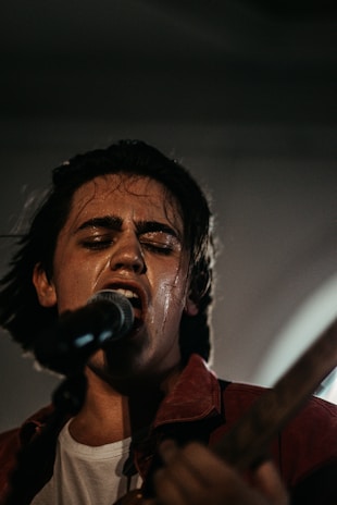 A person passionately singing into a microphone with closed eyes, implying deep emotional engagement. The setting appears dimly lit, creating a moody atmosphere. The individual is wearing a dark shirt and is slightly sweaty, suggesting a dynamic performance. The focus is primarily on the singer while the background is blurred.